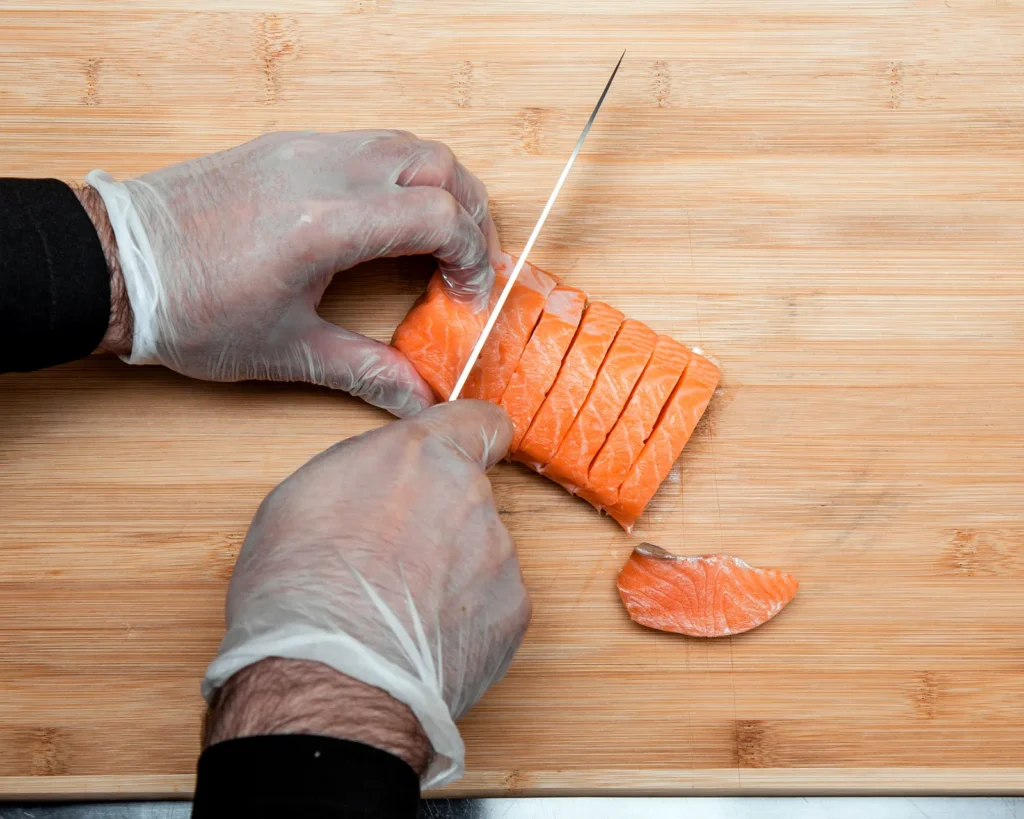 Mani con guanti trasparenti che tagliano un filetto di salmone crudo su un tagliere di legno