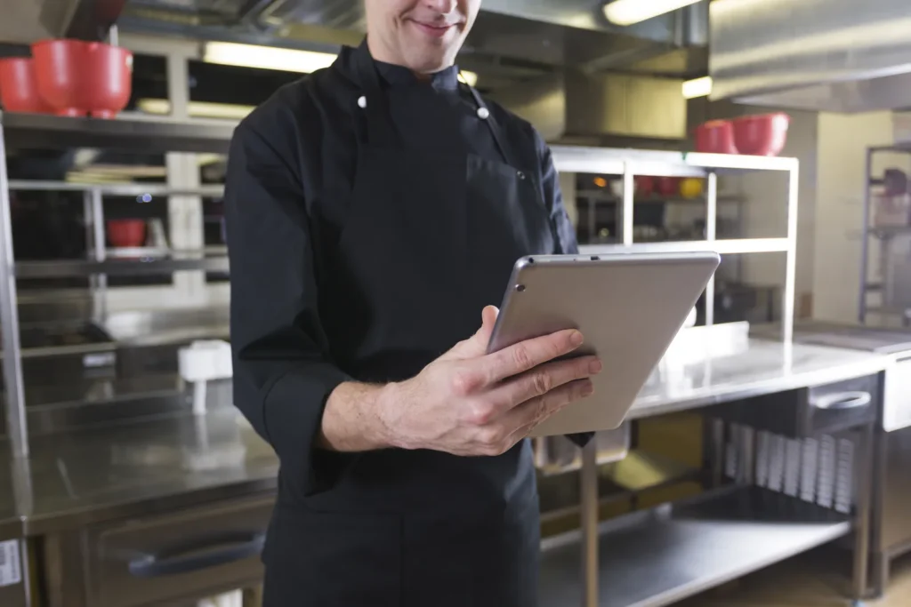 Cuoco in giacca nera che consulta un tablet in una cucina professionale in acciaio