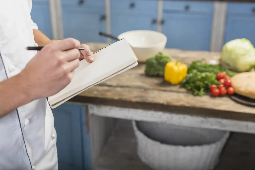 Cuoco in giacca bianca che prende appunti su un blocco in cucina accanto alle verdure